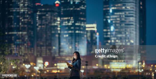 young businesswoman using digital tablet in financial district, against illuminated corporate skyscrapers at night - big tech foto e immagini stock