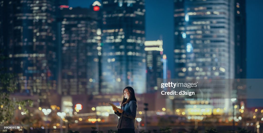 Young businesswoman using digital tablet in financial district, against illuminated corporate skyscrapers at night