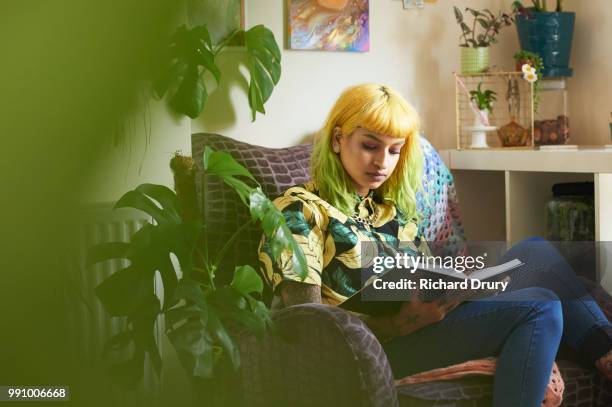 young hipster woman reading a book in her living room - room plant stockfoto's en -beelden