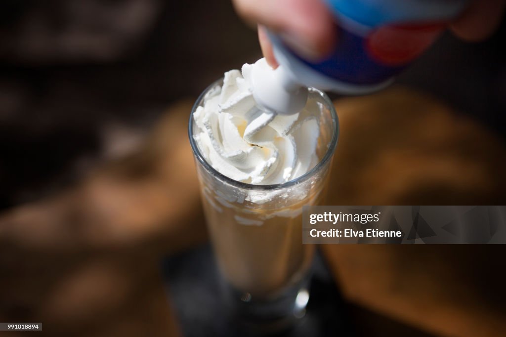 Whipped cream being sprayed into a tall glass of iced coffee