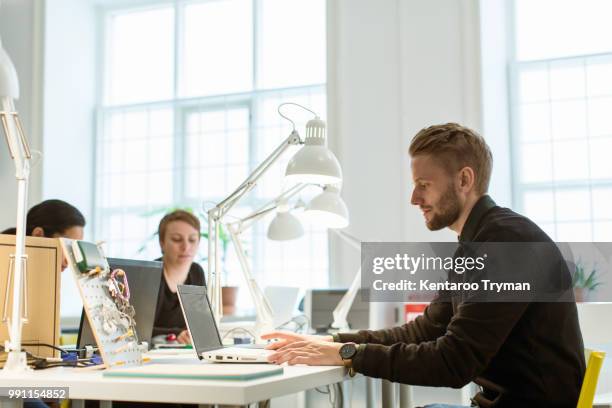 confident young businessman using laptop while sitting at desk in creative office - schreibtischlampe stock-fotos und bilder