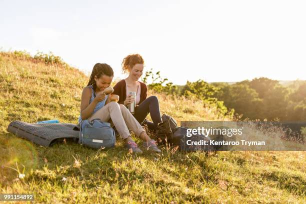 smiling woman and daughter with insulated drink container sitting on grass at hill against sky during sunset - insulated drink container foto e immagini stock
