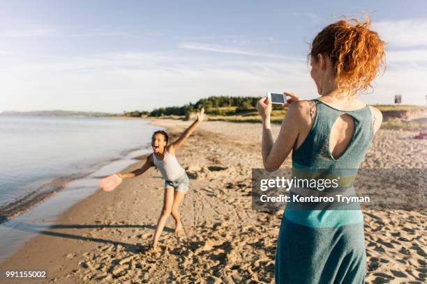 rear view of woman photographing playful daughter standing on shore at beach against sky - summer pictures stock pictures, royalty-free photos & images
