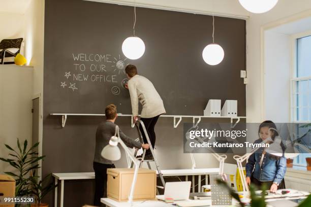 businesswoman standing on step ladder drawing smiley face on wall while colleagues working at creative office - smiley face stock pictures, royalty-free photos & images