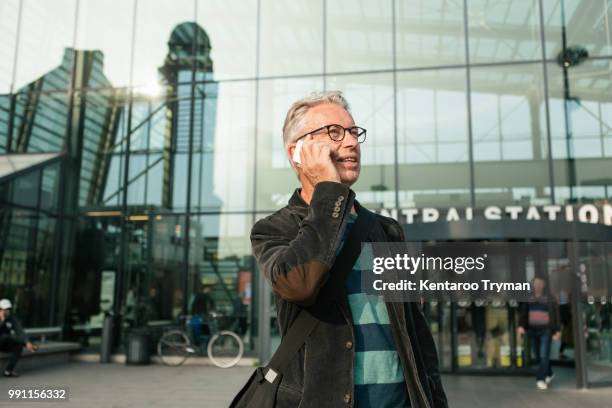 senior male commuter looking away while talking through smart phone against railroad station building - modern city life stock pictures, royalty-free photos & images