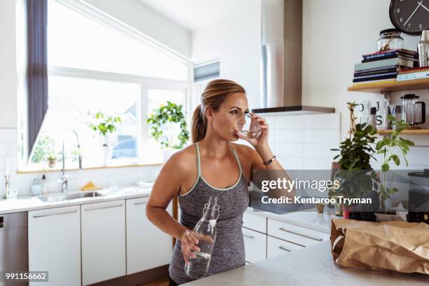 tired woman in sports clothing drinking water while standing at kitchen - women with health faucet stock pictures, royalty-free photos & images