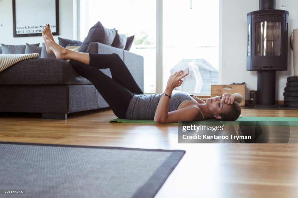 Tired woman using mobile phone while lying on exercise mat in living room