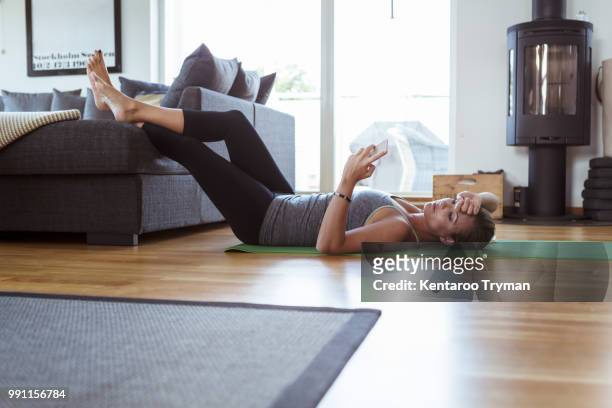 tired woman using mobile phone while lying on exercise mat in living room - feet on table bildbanksfoton och bilder
