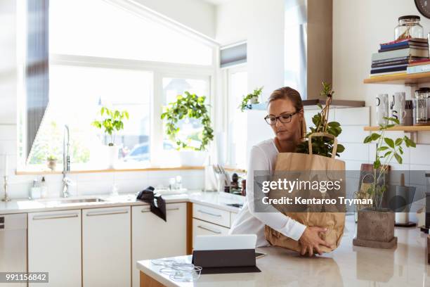 woman holding paper bag while looking at digital tablet in kitchen - frau tüte einkaufen stock-fotos und bilder