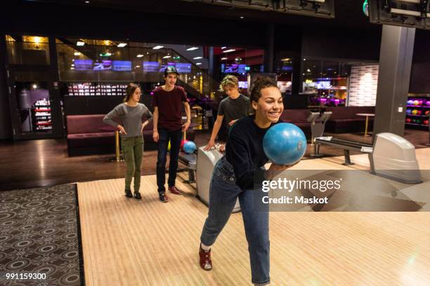 smiling teenage girl holding ball against friends standing on illuminated parquet floor at bowling alley - bowlingbahn stock-fotos und bilder