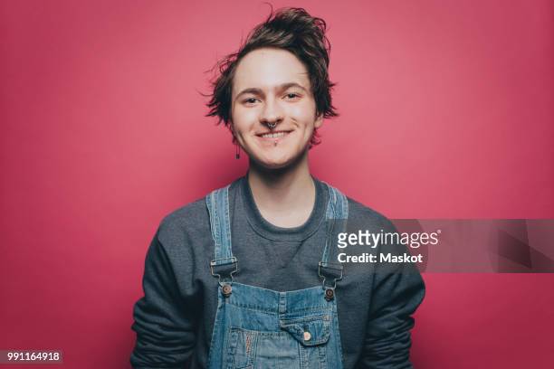 portrait of smiling young man wearing denim overalls over pink background - androgino fotografías e imágenes de stock