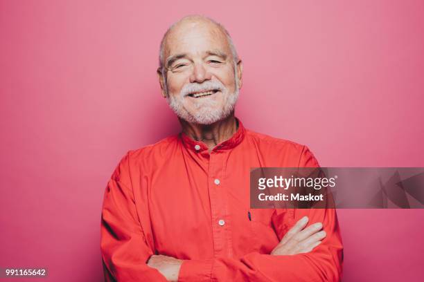 portrait of smiling senior man with arms crossed against pink background - mode et couleur photos et images de collection