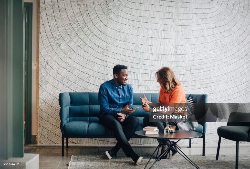 Businessman and woman taking while sitting on couch against wall at conference