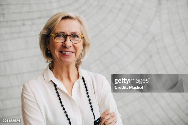 portrait of smiling senior businesswoman standing against wall in office - 60 64 years stock pictures, royalty-free photos & images