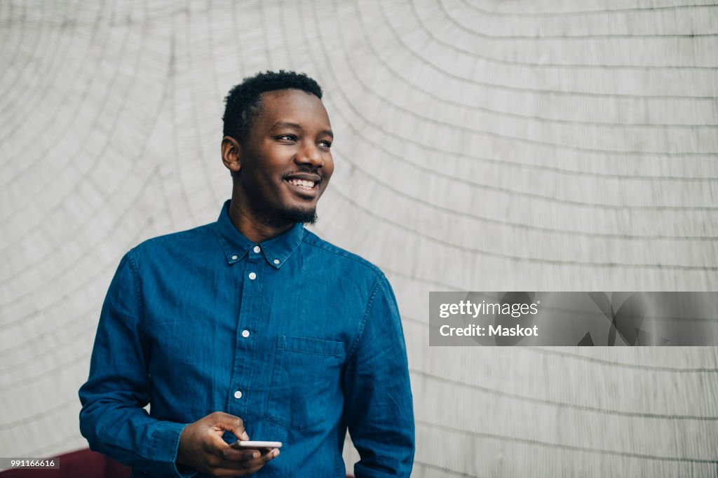 Smiling businessman holding mobile phone while looking away against gray wall
