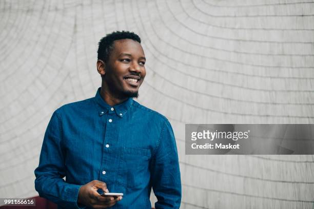 smiling businessman holding mobile phone while looking away against gray wall - informeel zakelijk stockfoto's en -beelden