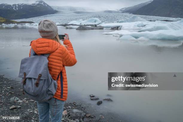 young woman taking mobile phone picture of glacier lake, icebergs floating on water - south east iceland stock pictures, royalty-free photos & images