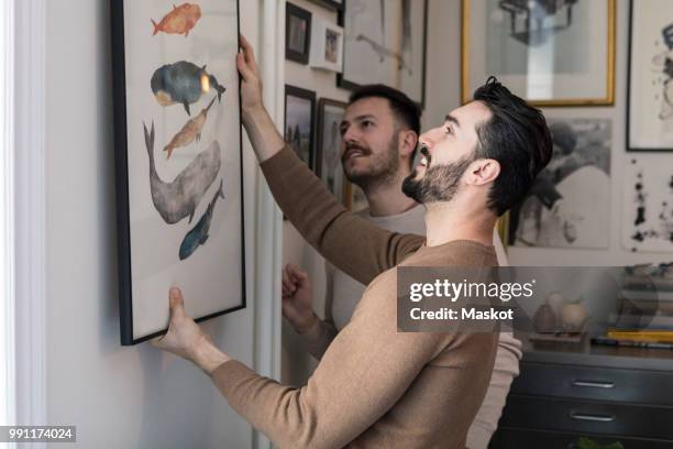 homosexual couple hanging painting on wall at home - decorating photos et images de collection