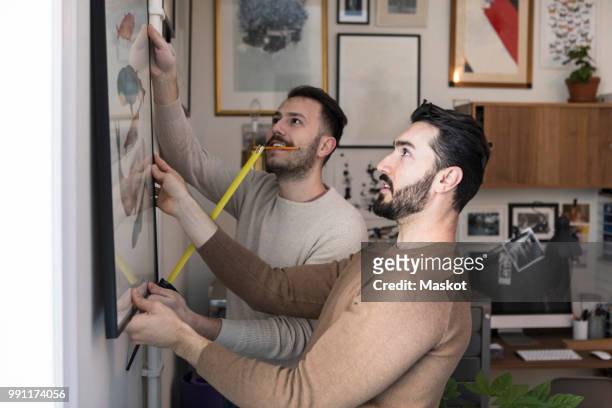 young man with tape measure assisting partner while hanging painting on wall - house for an art lover stock pictures, royalty-free photos & images
