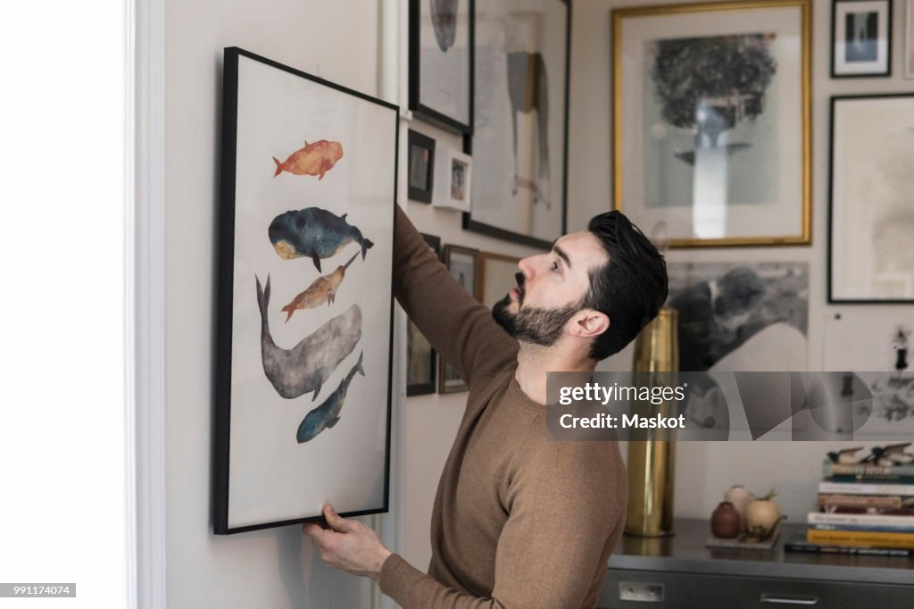 Young man hanging painting on wall at home