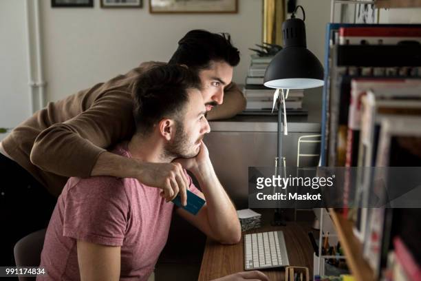 Gay couple shopping online through credit card on computer at illuminated desk in living room
