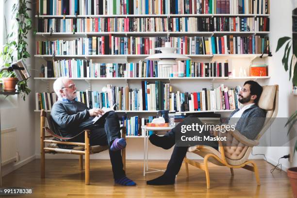 therapist communicating with man while sitting by book shelf at home office - counseling stockfoto's en -beelden