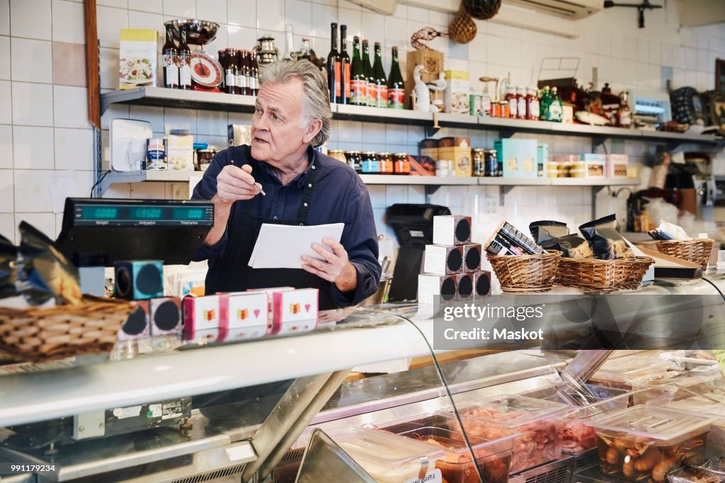 Male owner looking away while holding pen and note pad at counter in store