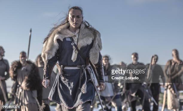 een individuele vrouwelijke viking poseren voor een groep van krijgers stond in de branding op de kust - larp stockfoto's en -beelden