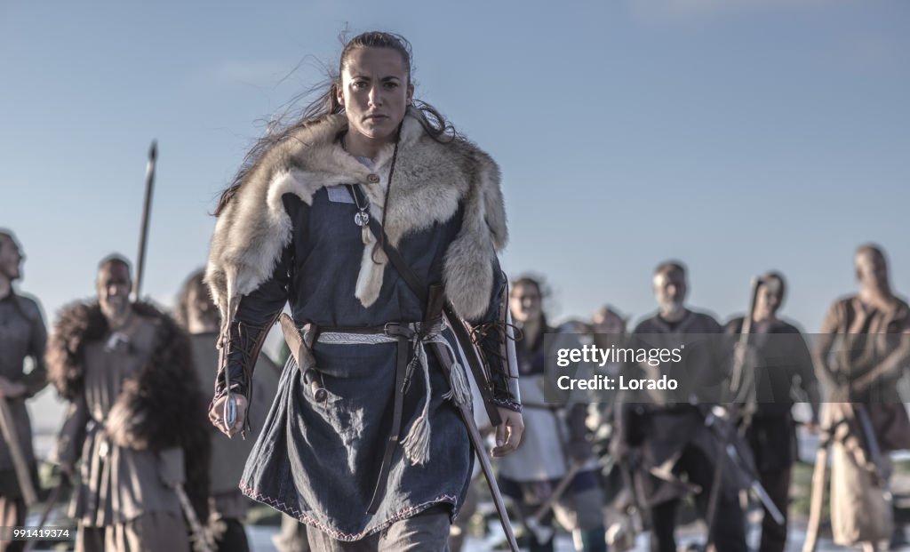 An individual female viking posing in front of a group of warriors stood in the surf on the shore