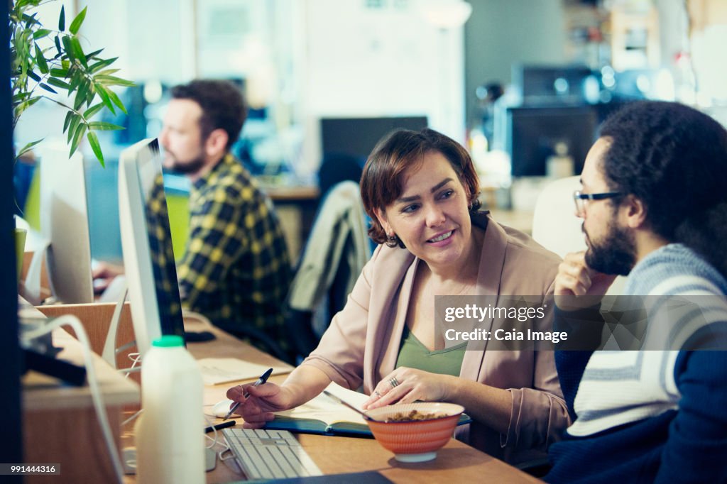 Creative business people eating cereal, meeting at computer in open plan office