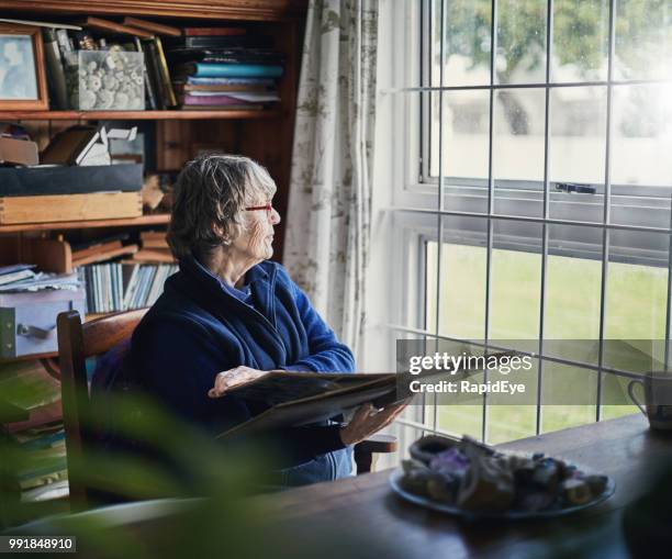 senior woman holding photo album looks through window - grass scrapbook stock pictures, royalty-free photos & images