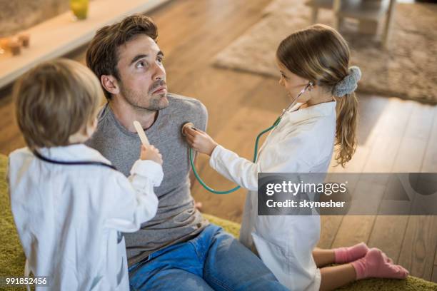 small siblings having fun while giving their father medical exam at home. - adult imitation stock pictures, royalty-free photos & images