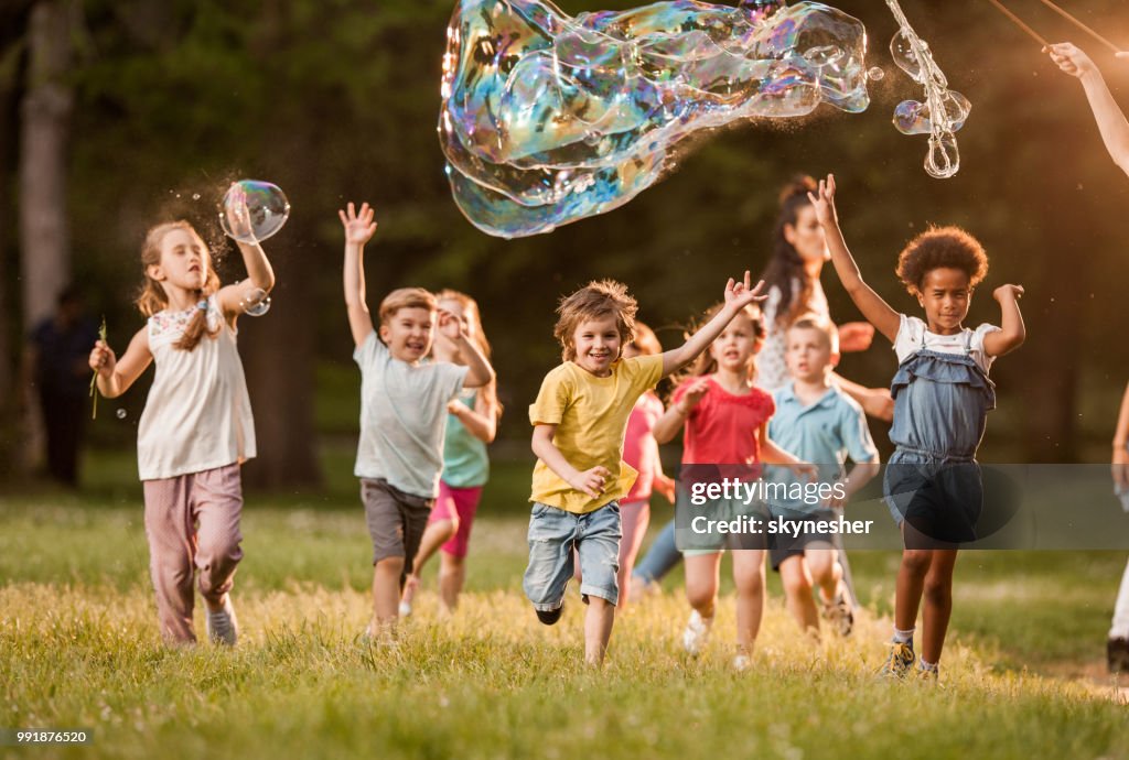 Playful kids having fun while running below rainbow bubbles in nature.