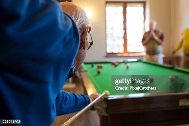 over shoulder focused and competitive active senior man lines up snooker cue and ball ready to take a shot - old pool table stock pictures, royalty-free photos & images
