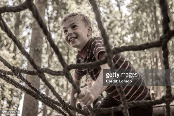 bambino maschio pre-adolescente individuale che si diverte sportivamente su un ponte di corda in un percorso ad ostacoli pubblico di corsa al fango - scala di corda foto e immagini stock