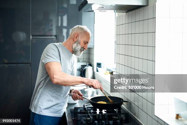 senior man with grey beard using frying pan in modern kitchen - cuisiner photos et images de collection