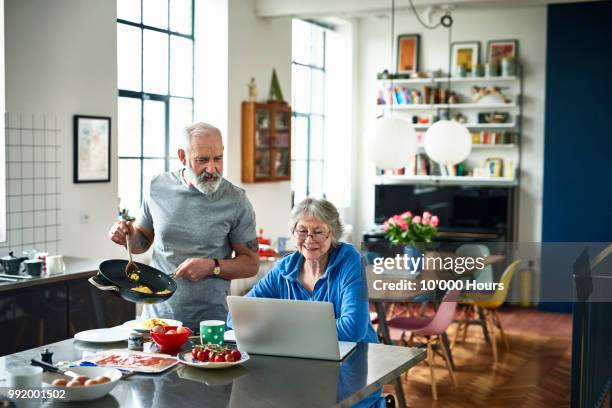 senior woman using laptop and smiling as man serves breakfast - couple breakfast stock-fotos und bilder