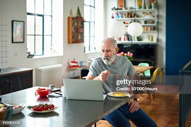 senior man sitting in kitchen eating breakfast and looking at laptop - old person kitchen food stock-fotos und bilder