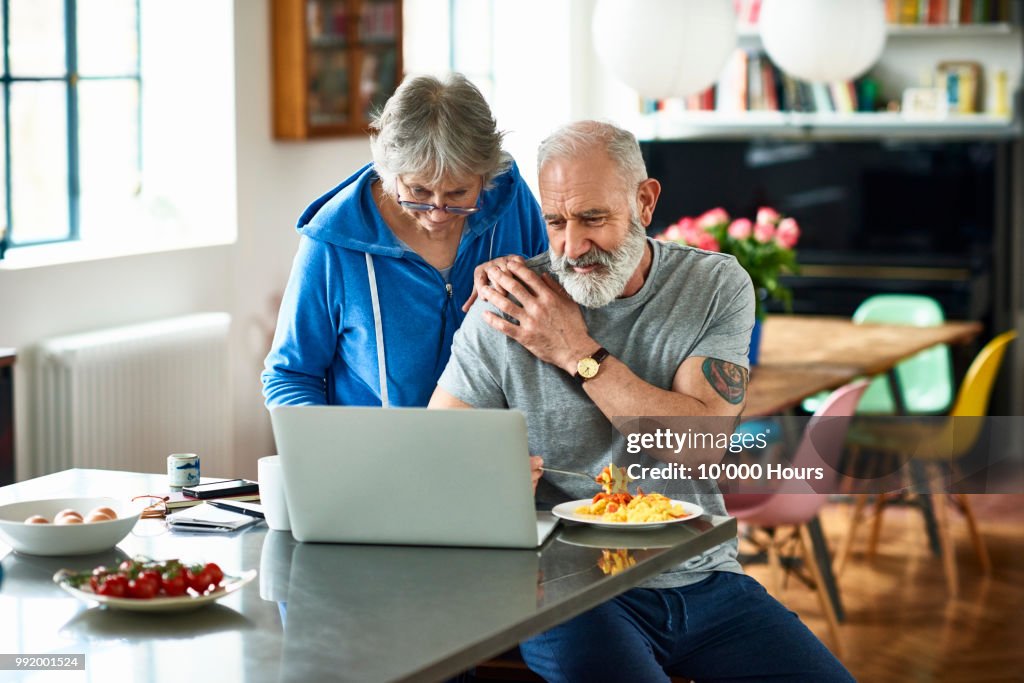 Affectionate retired couple with laptop in kitchen