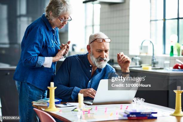 senior man using laptop and woman watching over shoulder - hours around the world stock pictures, royalty-free photos & images
