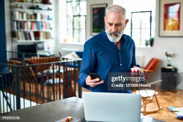 senior creative professional remote working and checking phone at home - man middelbare leeftijd woonkamer stockfoto's en -beelden