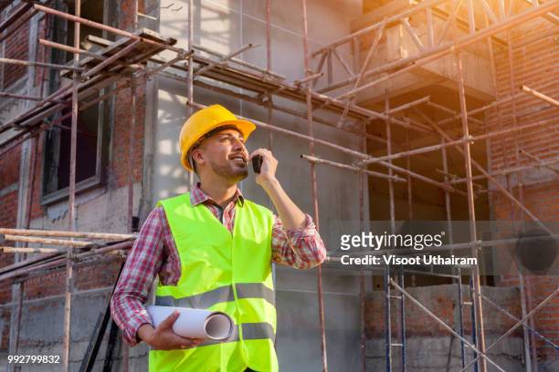 confident supervisor in hard hat holding blueprint and talking on the mobile phone while standing outdoors - business man overseeing blueprints outdoor stock-fotos und bilder