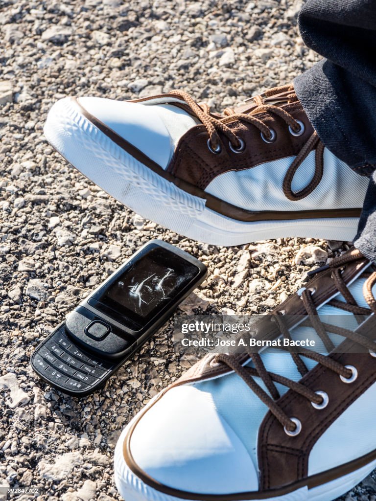 Slipper of white color of a person squishing a mobile phone against the soil, outdoors.