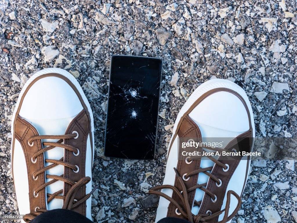 Slipper of white color of a person squishing a mobile phone against the soil, outdoors.