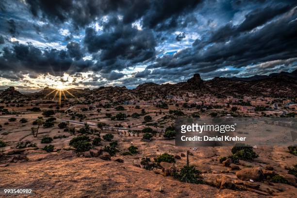 small village of aguard oudad with brightly painted houses in front of the rock chapeau napoleon, dramatic clouds, evening, tafraoute, anti atlas, southern morocco, morocco - chapeau ストックフォトと画像