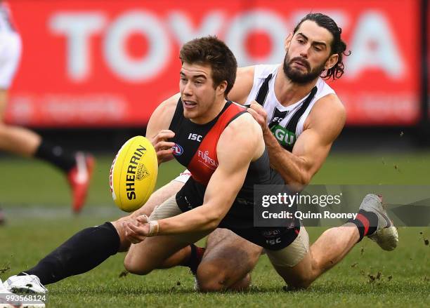 Zach Merrett of the Bombers handballs whilst being tackled by Brodie Grundy of the Magpies during the round 16 AFL match between the Essendon Bombers...