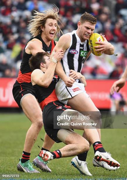 Taylor Adams of the Magpies is tackled by Zach Merrett and Dyson Heppell of the Bombers during the round 16 AFL match between the Essendon Bombers...