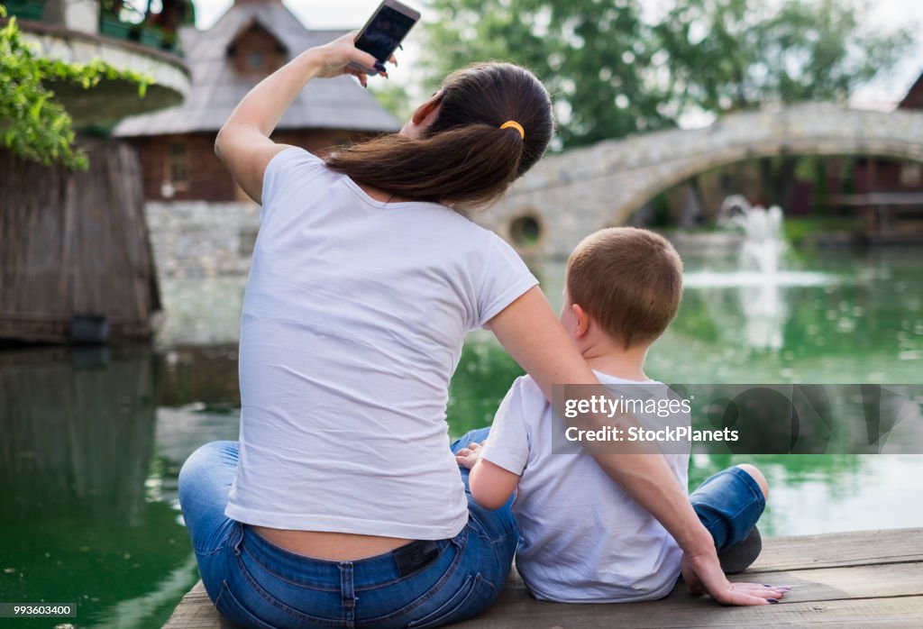 Back view mother taking a selfie with little son sitting near river