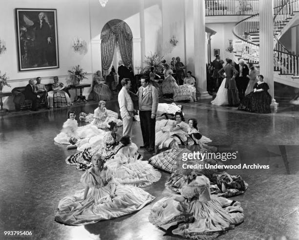 As female cast members in ball gowns sit on the floor, American assistant director Robert A Golden and director Richard Boleslawski discus a scene...