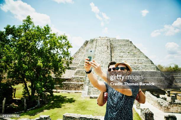 smiling gay couple taking selfie with smartphone while exploring mayapan ruins during vacation - cell mates stock-fotos und bilder